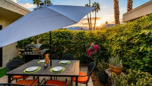 Une terrasse extérieure avec une table dressée pour les repas, un barbecue et un grand parasol pour faire de l’ombre.