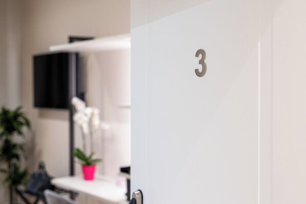 A room with a white door marked with the number 3, a white table, a pink plant, and a black shelf with a flat-screen TV. - Relais Cavallini (Rome)