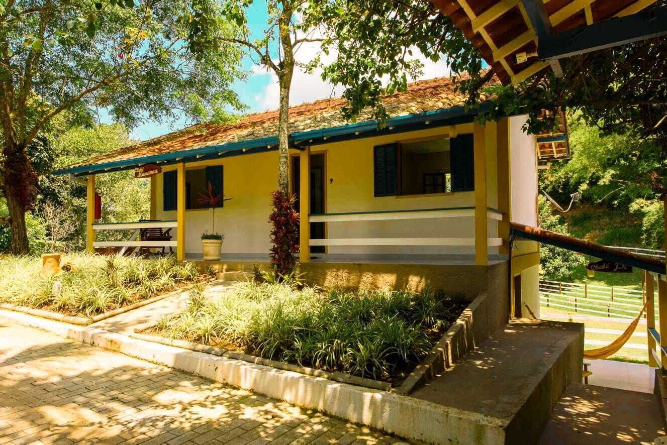 A yellow building with a tiled roof, surrounded by greenery and a paved area.