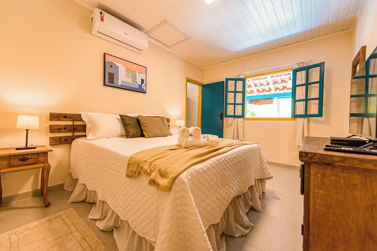 Une chambre à coucher comprenant un lit, une table de chevet, une commode, une fenêtre avec des volets bleus et un appareil de climatisation.