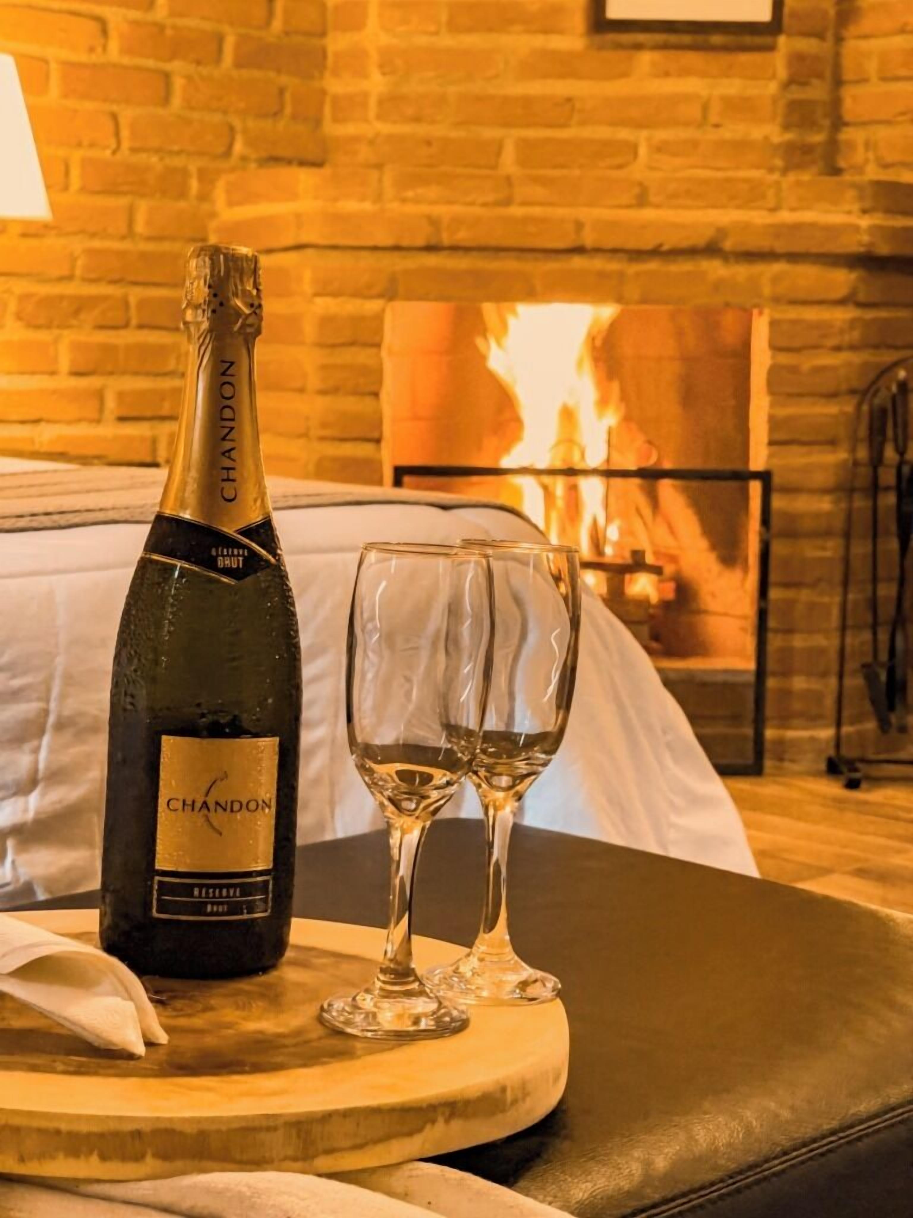 A bottle of Chandon Brut champagne and two glasses on a wooden tray in a hotel room with a brick wall and a fireplace.