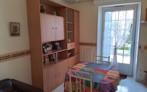 A room with a wooden cabinet, a table with a colorful quilt, a chair, a radiator, and a window with a polka dot curtain. - Holiday Home 'La Maison Bleue' with Private Terrace, Private Garden and Wi-Fi (Thésée)