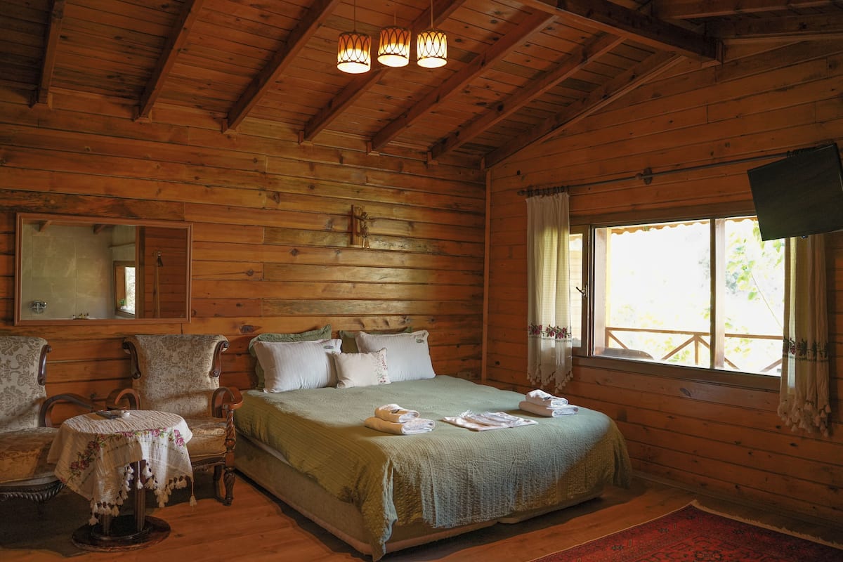 A wooden cabin bedroom with a bed, a small table, a mirror, and a window.