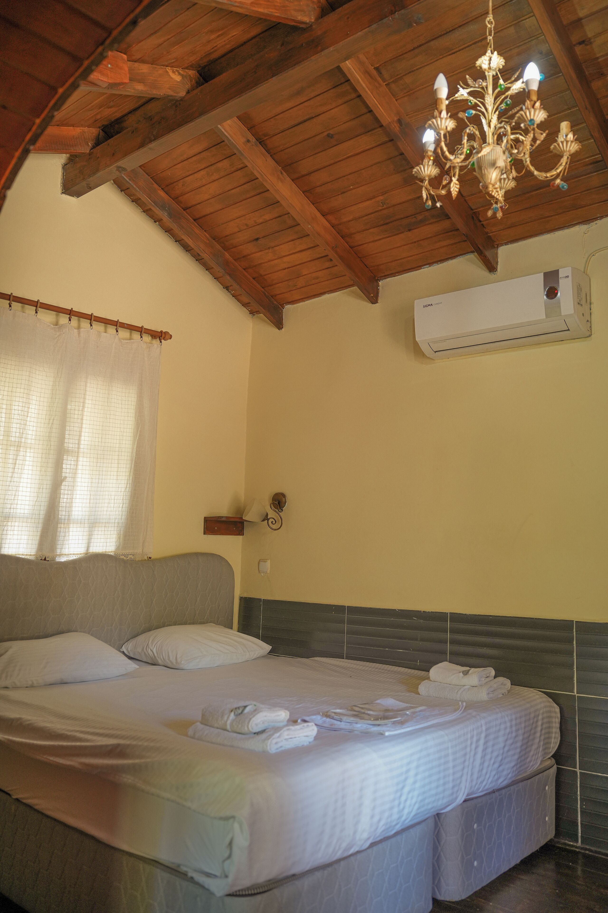 A bedroom with a wooden ceiling, a chandelier, a bed with white linens, and an air conditioning unit on the wall.
