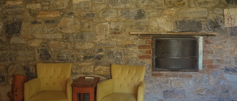 A room with a stone wall, two yellow armchairs, a small wooden table, and a fireplace with a metal grate.