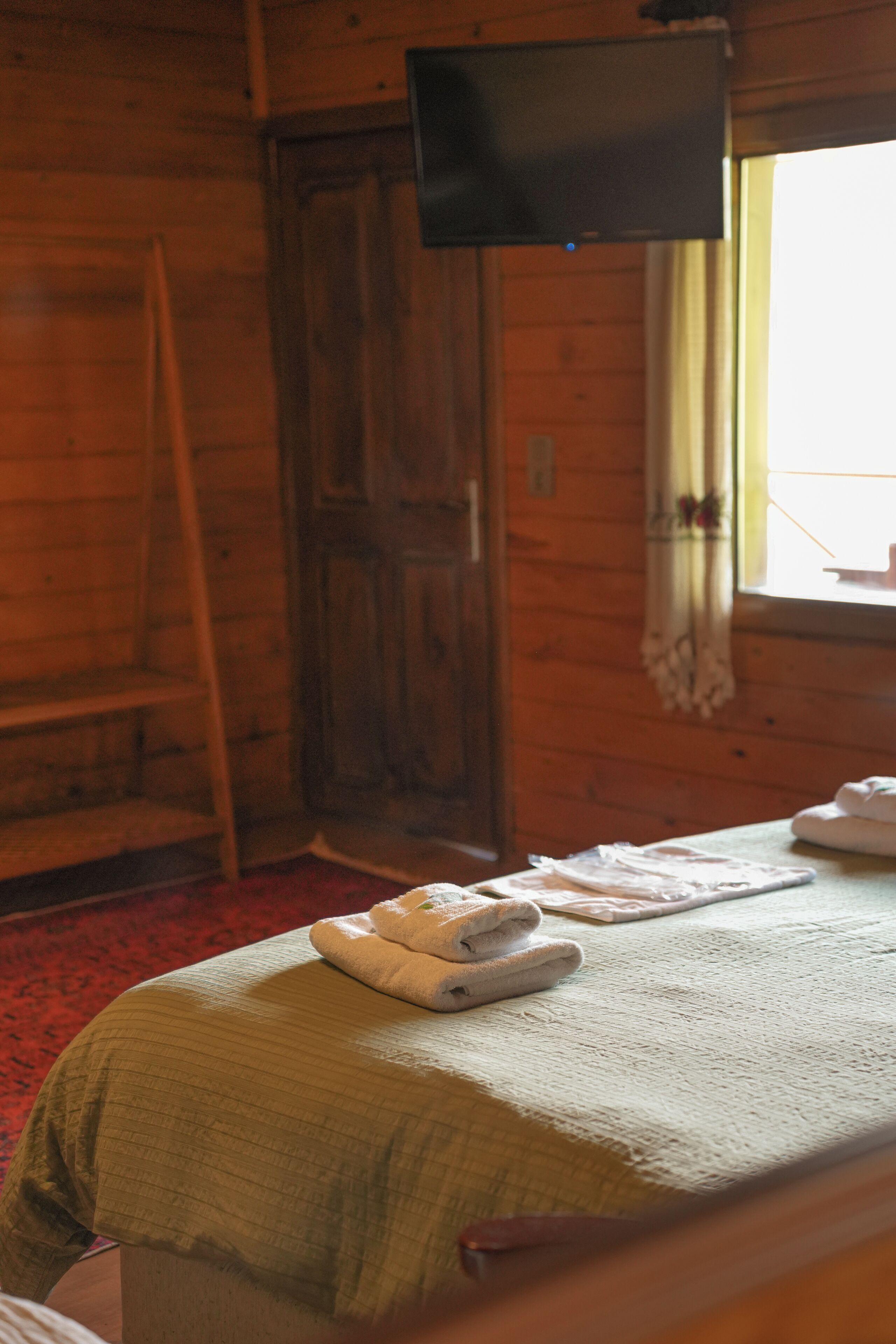 A wooden cabin room with a bed, a television mounted on the wall, and a window with curtains.