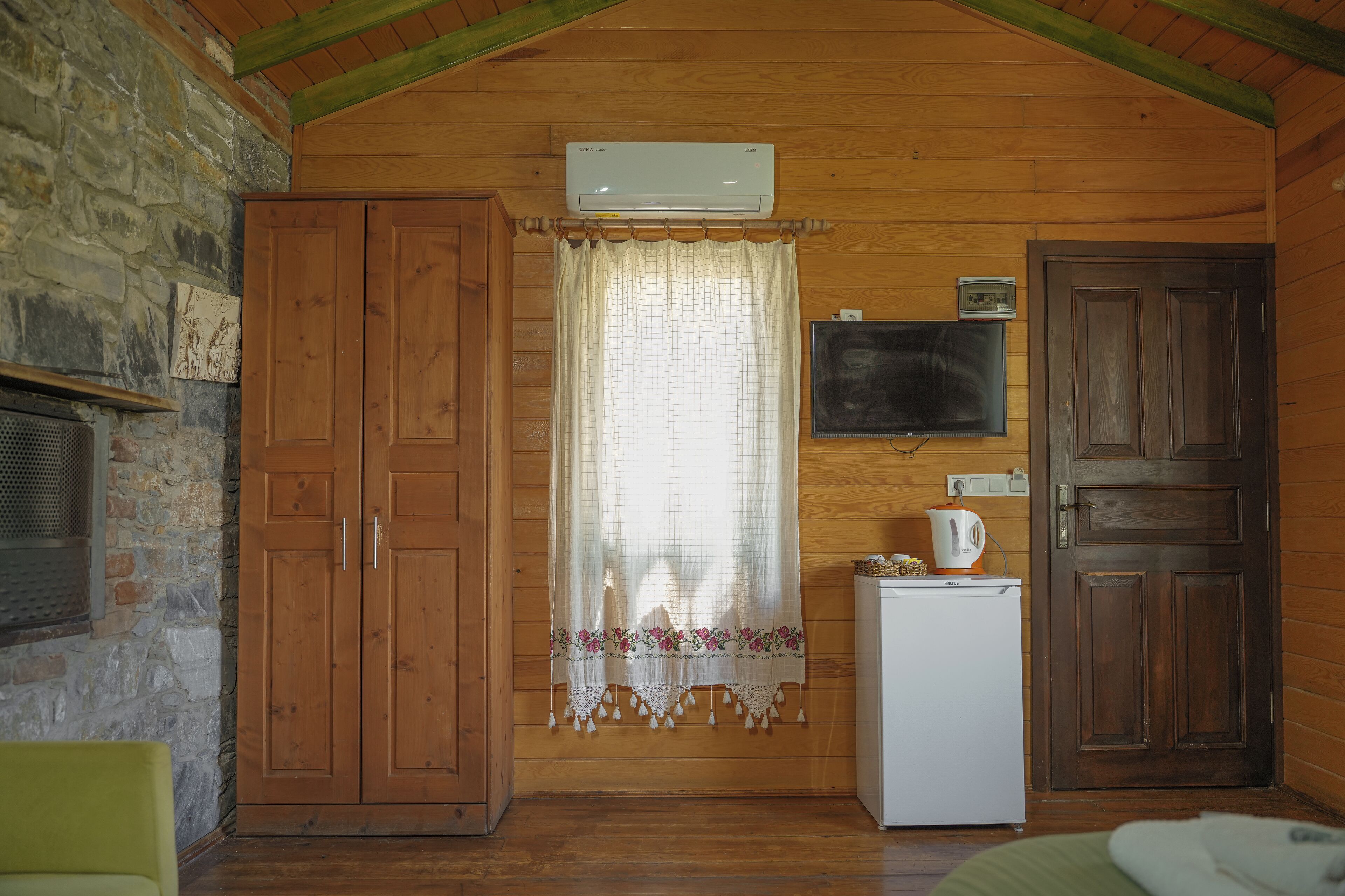 A cozy room with wooden walls, a stone fireplace, a wooden wardrobe, a small refrigerator, a television, and a door.