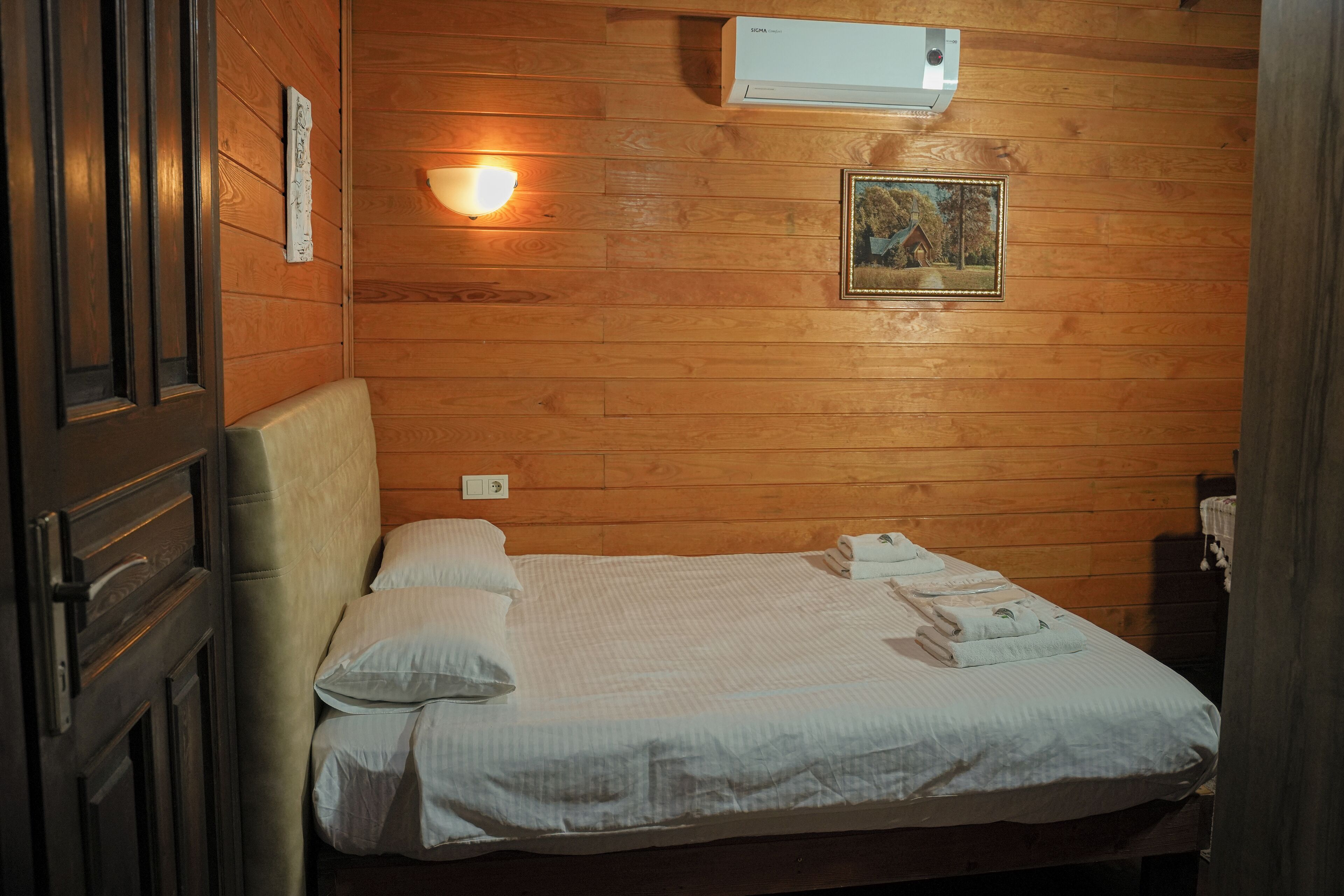 A wooden-paneled room with a bed, a wall-mounted air conditioner, a wall-mounted light, and a framed picture.