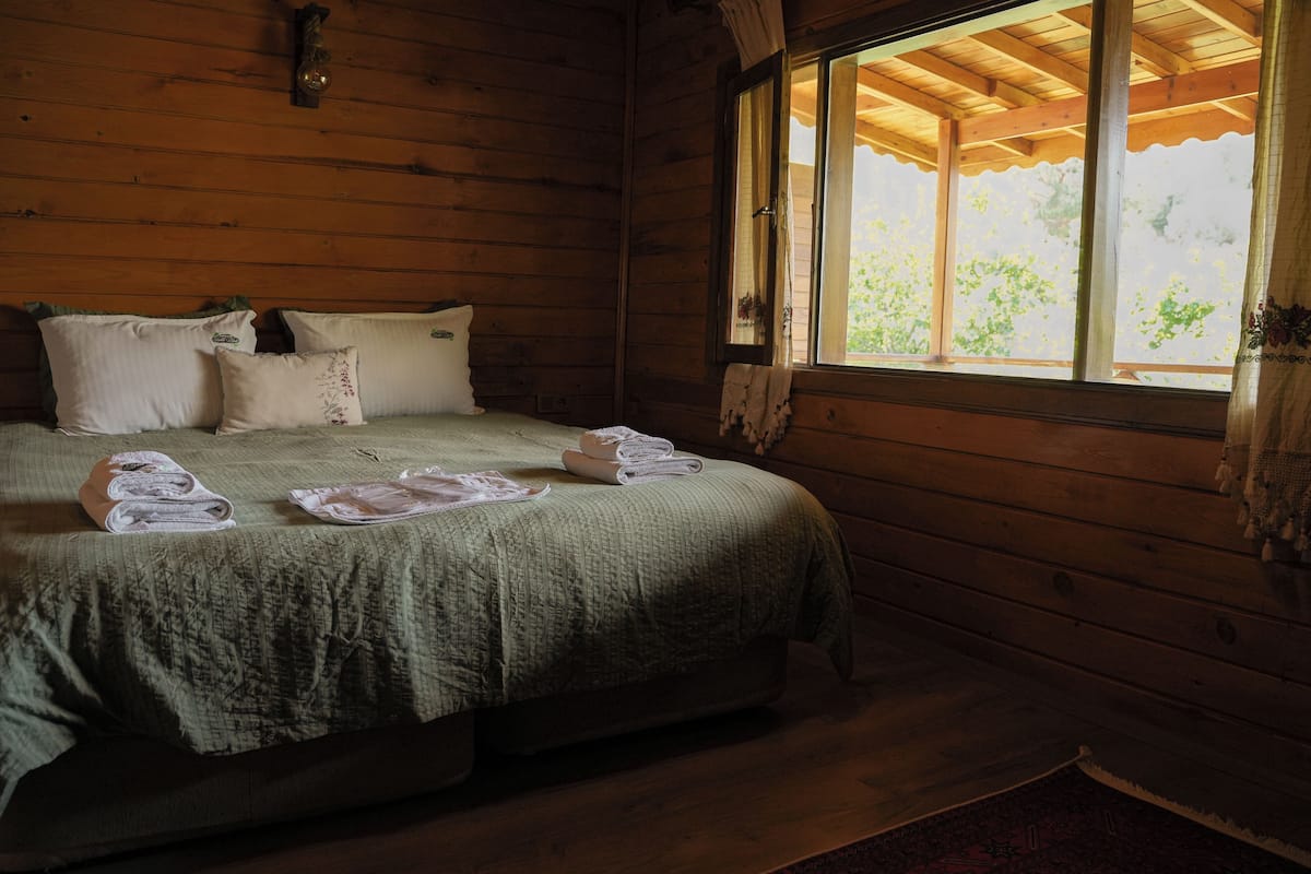 A wooden cabin room with a bed, pillows, and a window with a view of greenery.