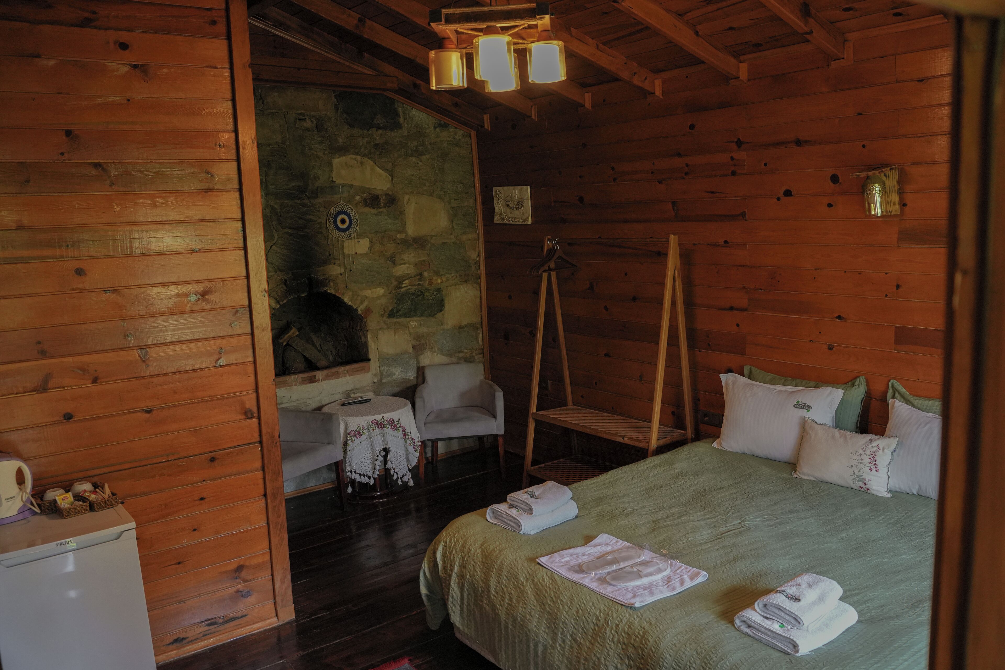 A wooden cabin room with a bed, a small table, a chair, and a stone wall.