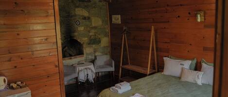 A wooden cabin room with a bed, a small table, a chair, and a stone wall.