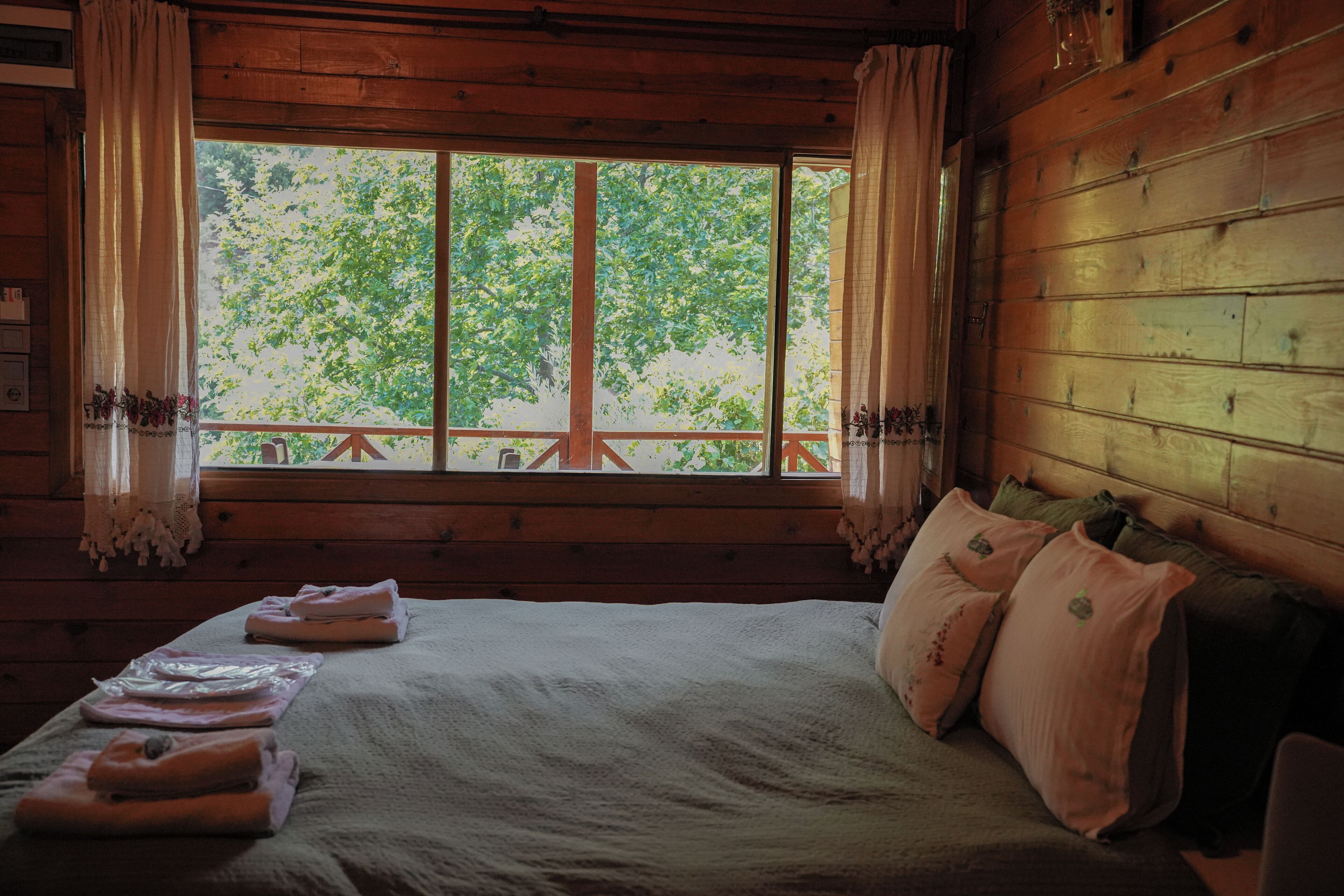 Habitación de cabaña de madera con una ventana grande, una cama con almohadas y vistas a árboles fuera.
