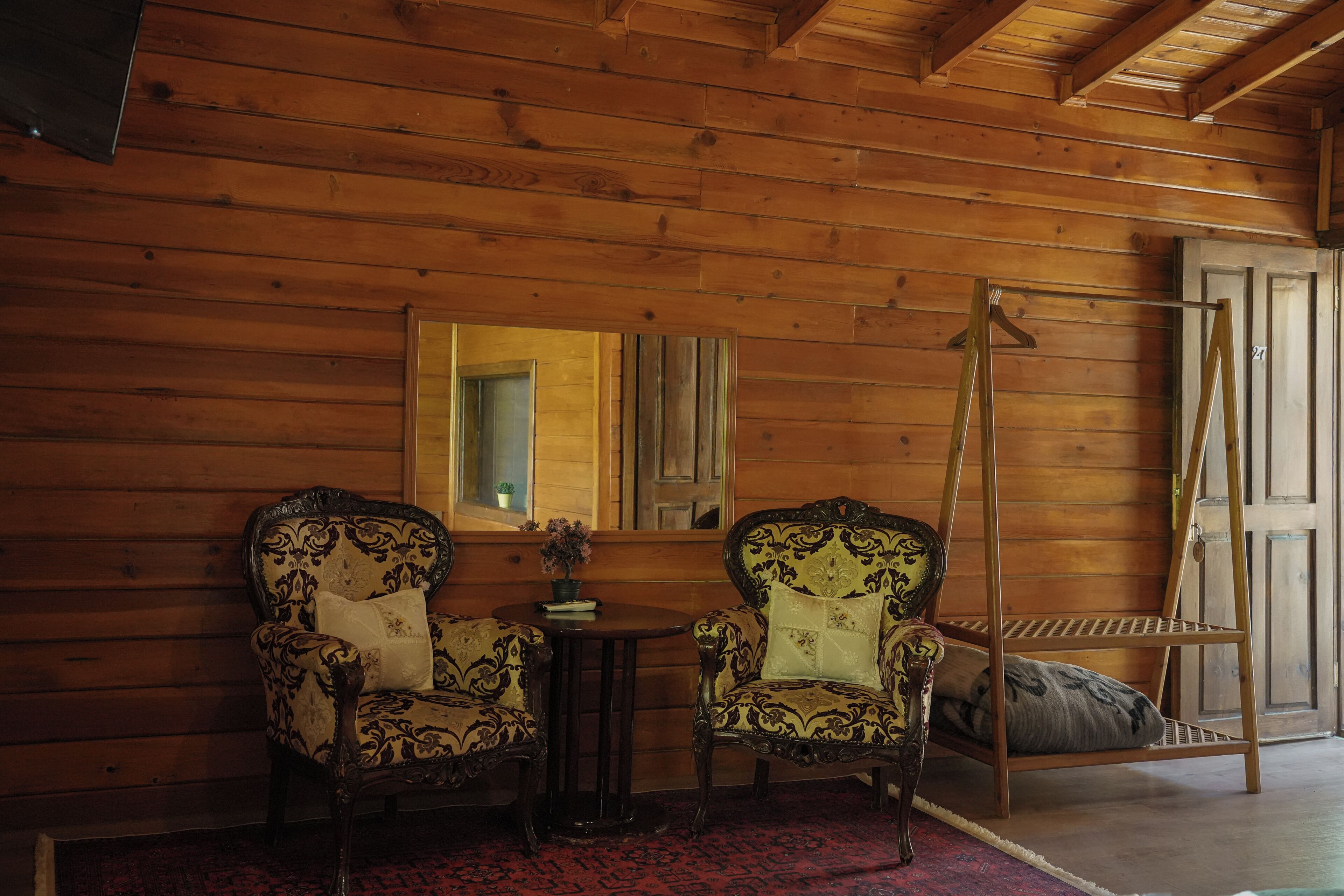 A wooden cabin interior with two ornate chairs, a small table with a plant, and a wooden swing.