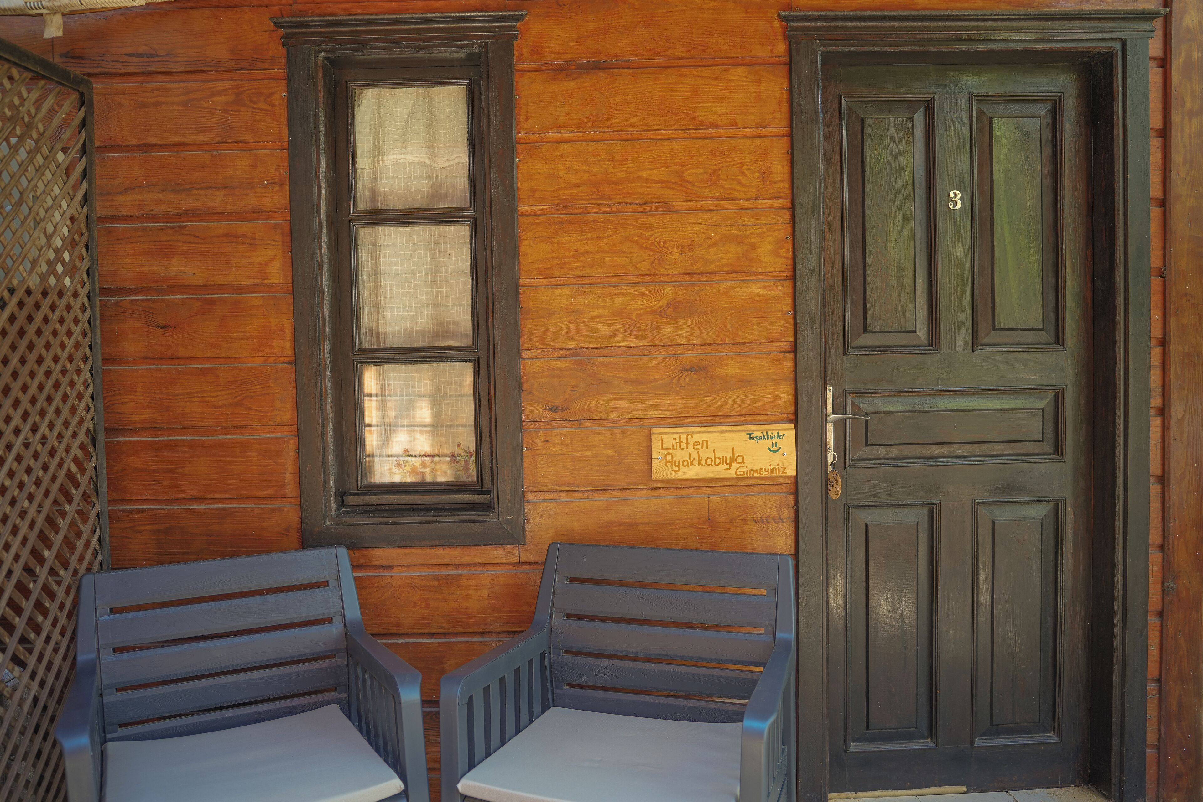 A wooden exterior wall with a dark door and a window. There is a sign above the door and two chairs in front of it.
