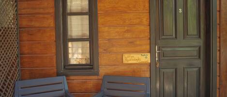 A wooden exterior wall with a dark door and a window. There is a sign above the door and two chairs in front of it.