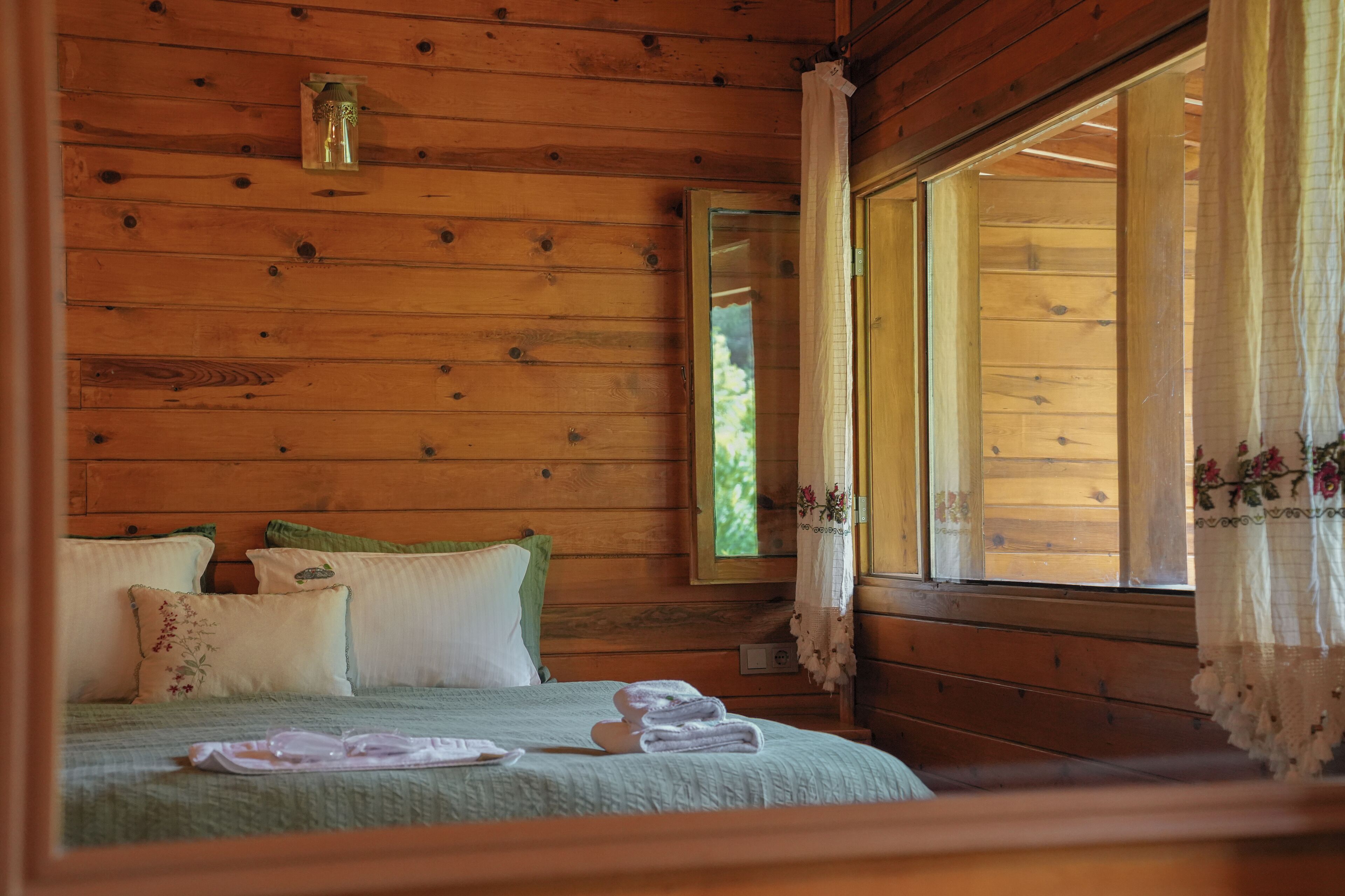 A wooden cabin bedroom with a bed, pillows, a nightstand, a window with curtains, and a mirror.