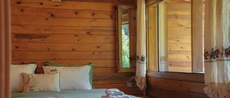 A wooden cabin bedroom with a bed, pillows, a nightstand, a window with curtains, and a mirror.