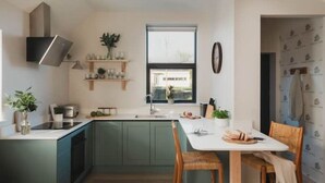A kitchen with green cabinets, a white countertop, a window, and a dining area with a table and chairs. - Longstones Avebury – Adam (Marlborough)