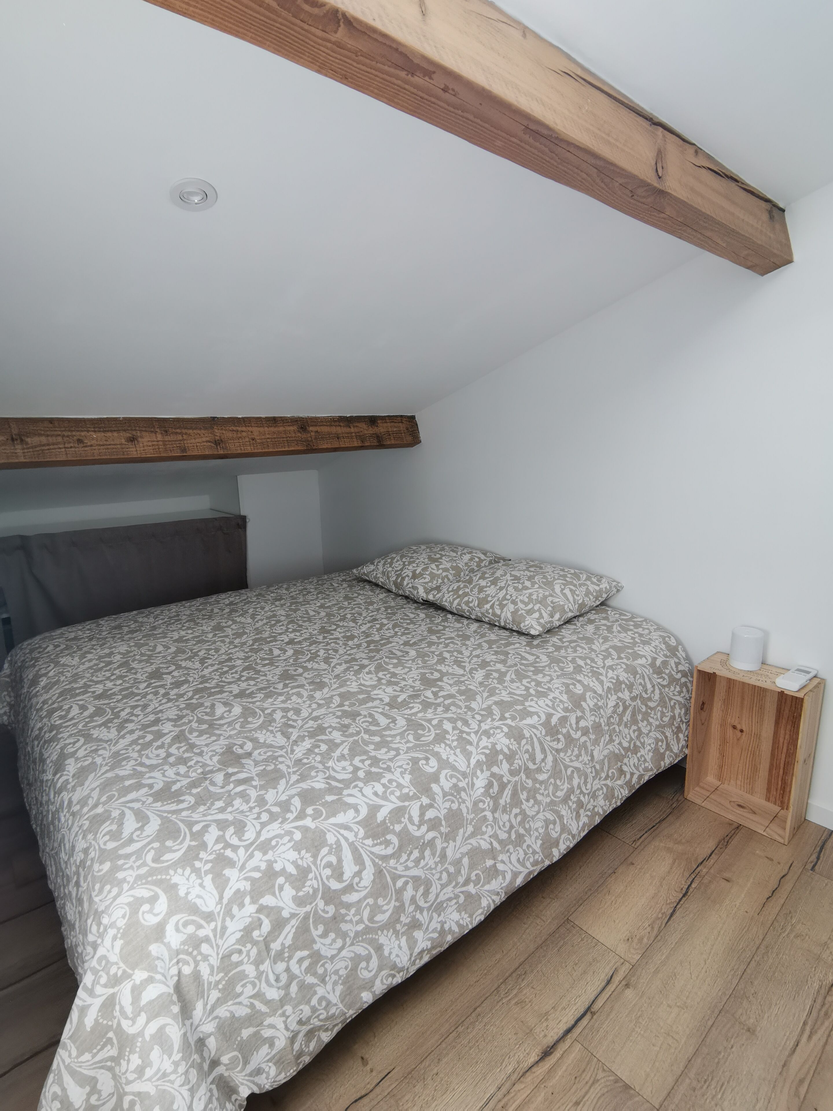 A bedroom with a bed, wooden beams, a bedside table with a lamp, and a wall-mounted light fixture.