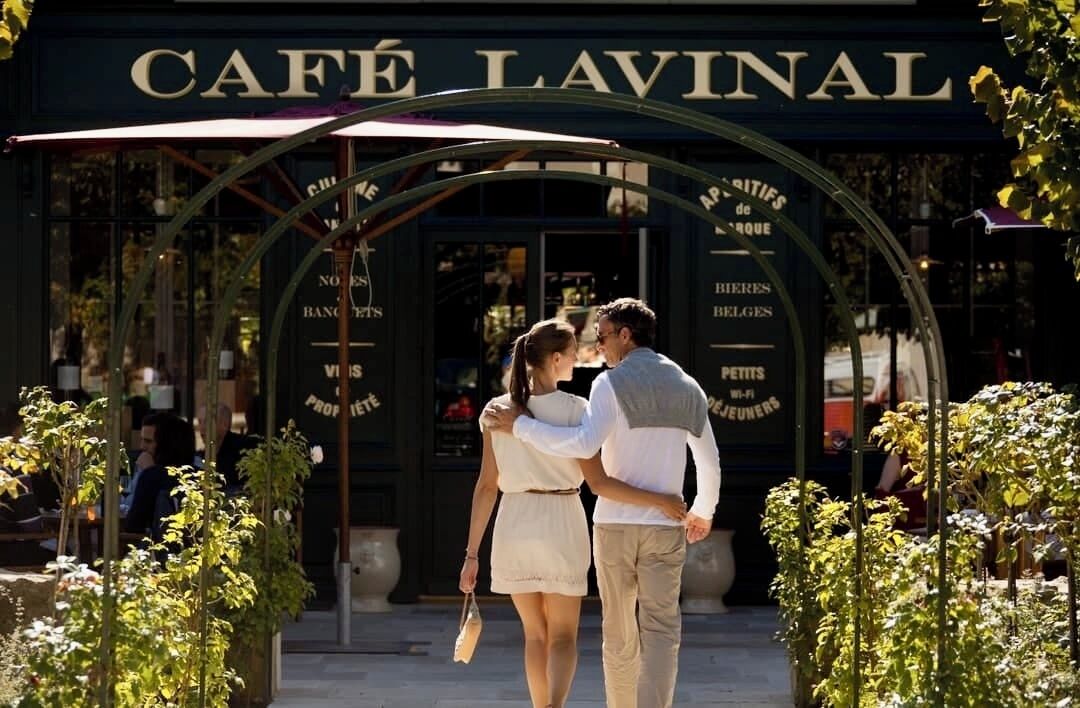 Een koppel loopt richting een café genaamd 'Café Lavinal' met een bord waarop menukaartitems staan vermeld zoals 'Noirs', 'Bangers', 'Vis', 'Petits', 'Wi-Fi', 'Belges' en 'Petits Jeuneurs'.