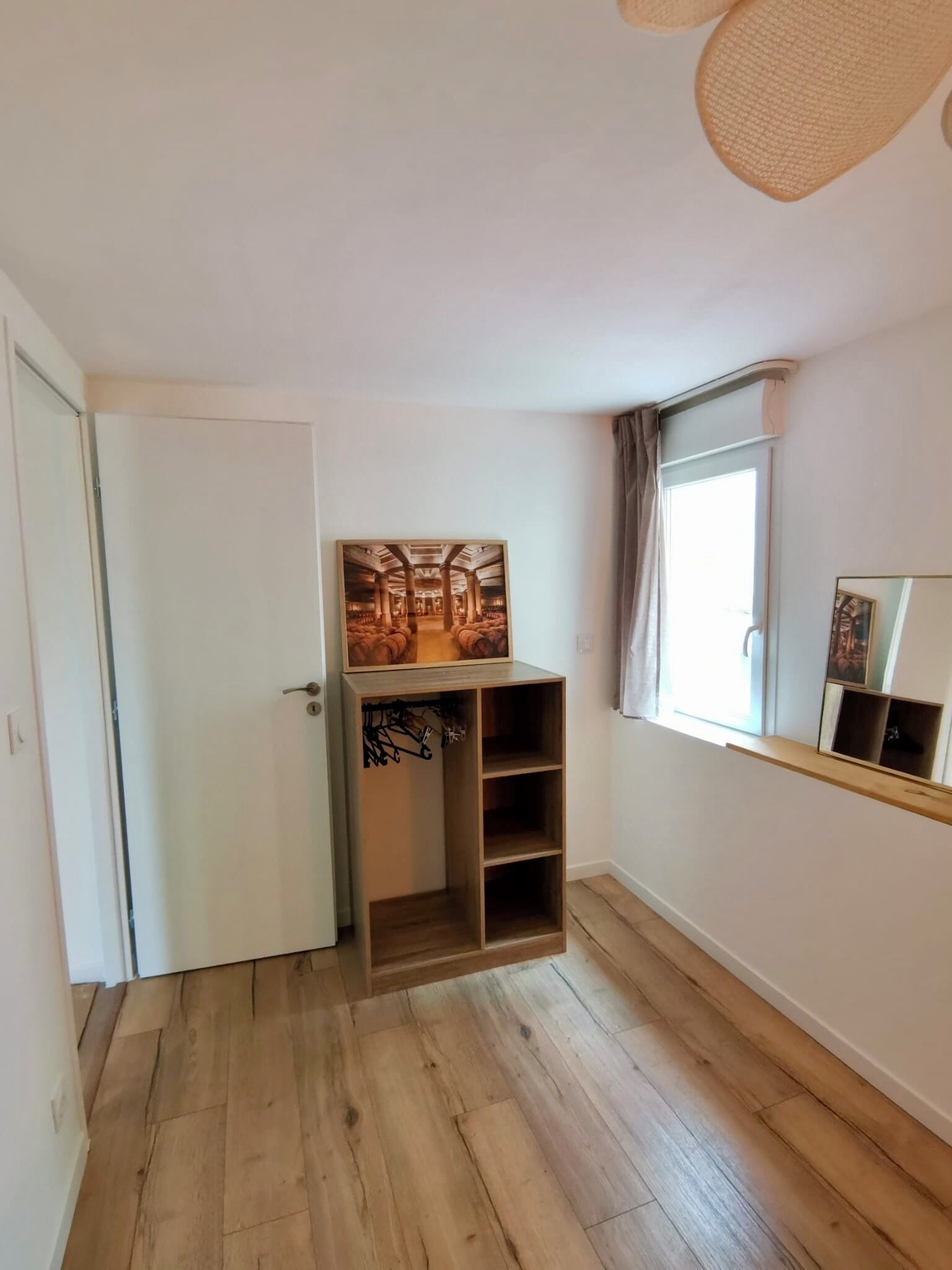 A room with wooden flooring, a white door, a built-in wooden shelf unit, and a window with curtains.