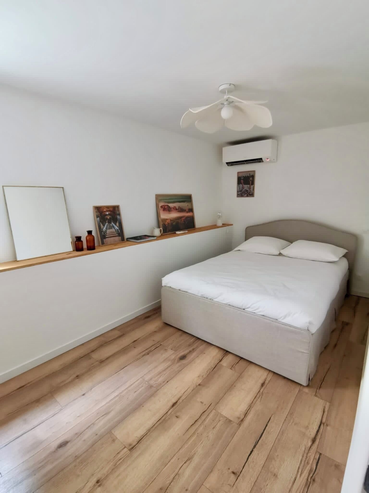 Une chambre à coucher comprenant un lit, un ventilateur de plafond, un appareil de climatisation, un plancher en bois et une étagère ornée d’objets décoratifs.