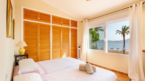 A bedroom with a bed, a nightstand, a lamp, a window with a view of the beach, and a wooden wardrobe. - Villa 'La Gola' with Sea Views, Shared Pool and Wi-Fi (La manga)