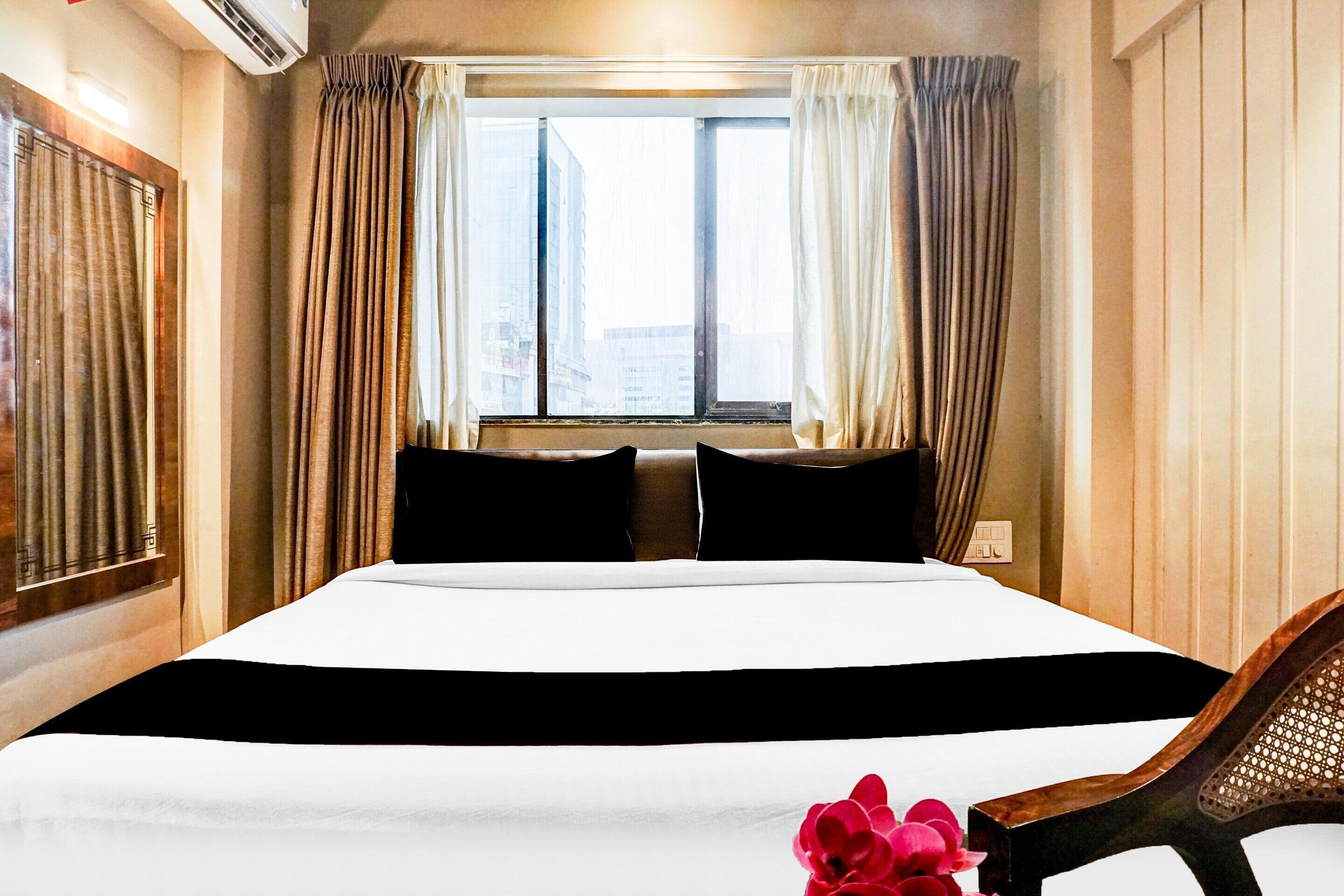 A hotel room with a large bed, a window with curtains, a wall-mounted air conditioner, and a wooden chair.