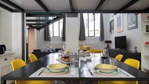 A modern dining area with a long table set for two, yellow chairs, and a television in the background. - Italianway Comfort - Mancini 3 (Milan)