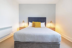 A neatly made bed with a grey duvet, a dark headboard, and a yellow throw pillow. There are two bedside tables with lamps, and a radiator is visible on the wall.