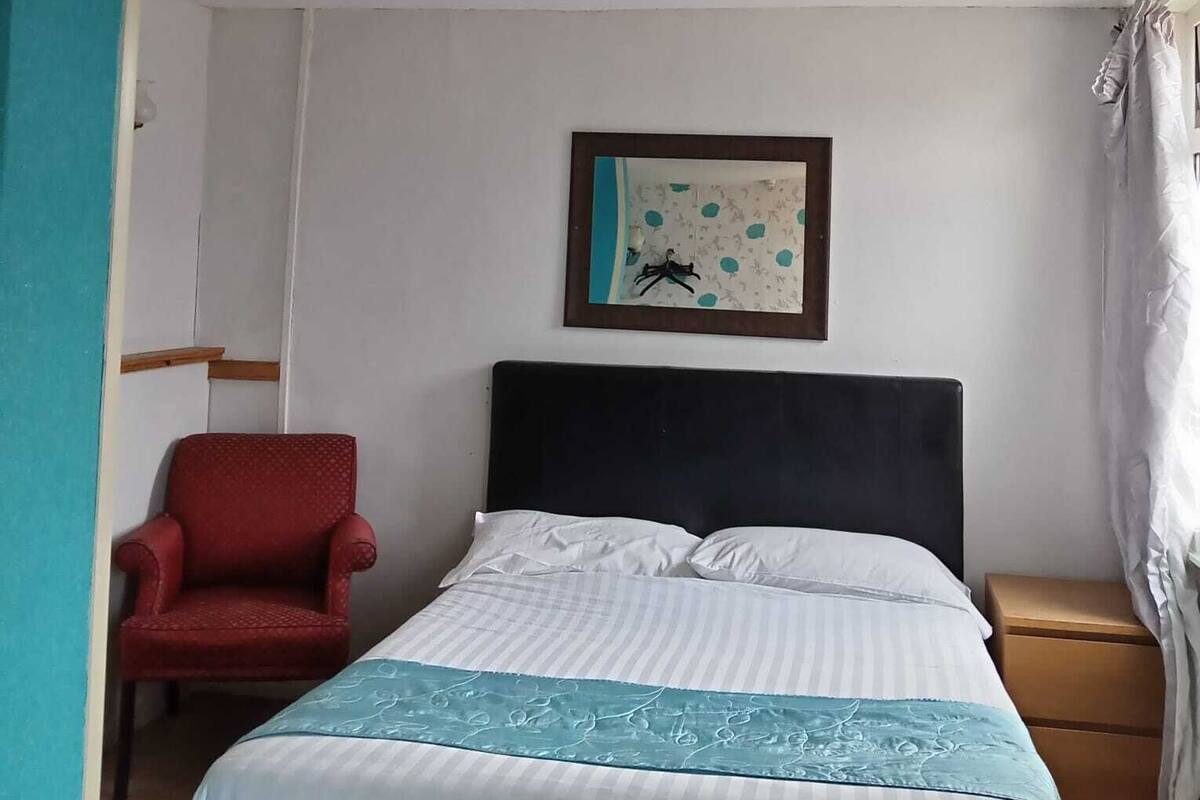 Un dormitorio con una cama, una silla roja, un cuadro en la pared, una lámpara en el techo y una ventana con cortinas.