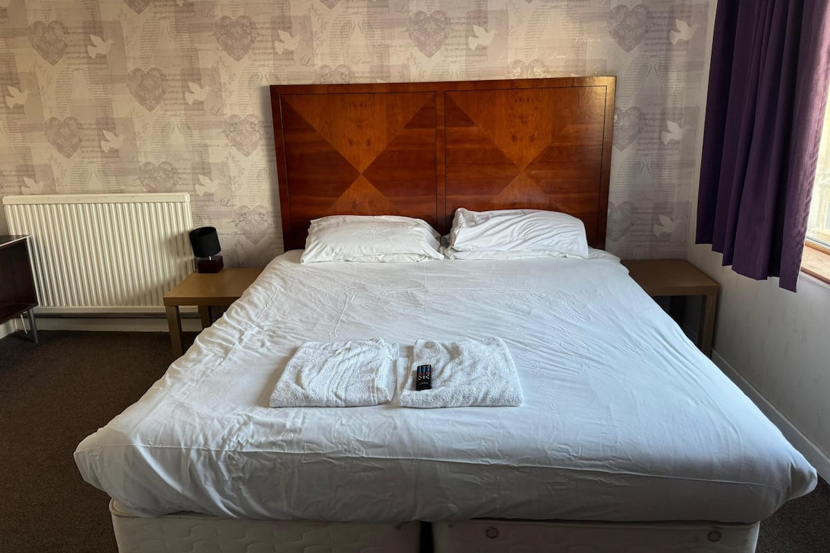 Una cama bien hecha con sábanas blancas, cabecera de madera y una mesita de noche con lámpara.