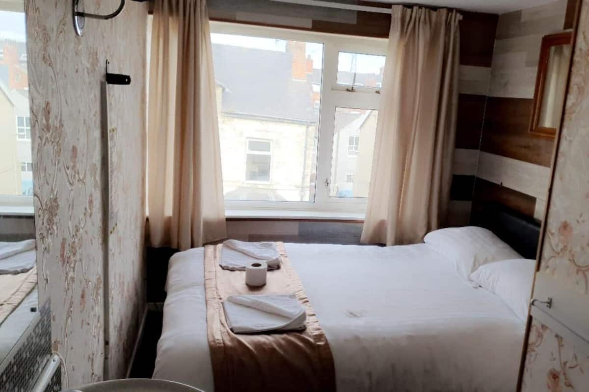 Habitación de hotel con cama, ventana con cortinas, cabecero de madera y vista a edificios.