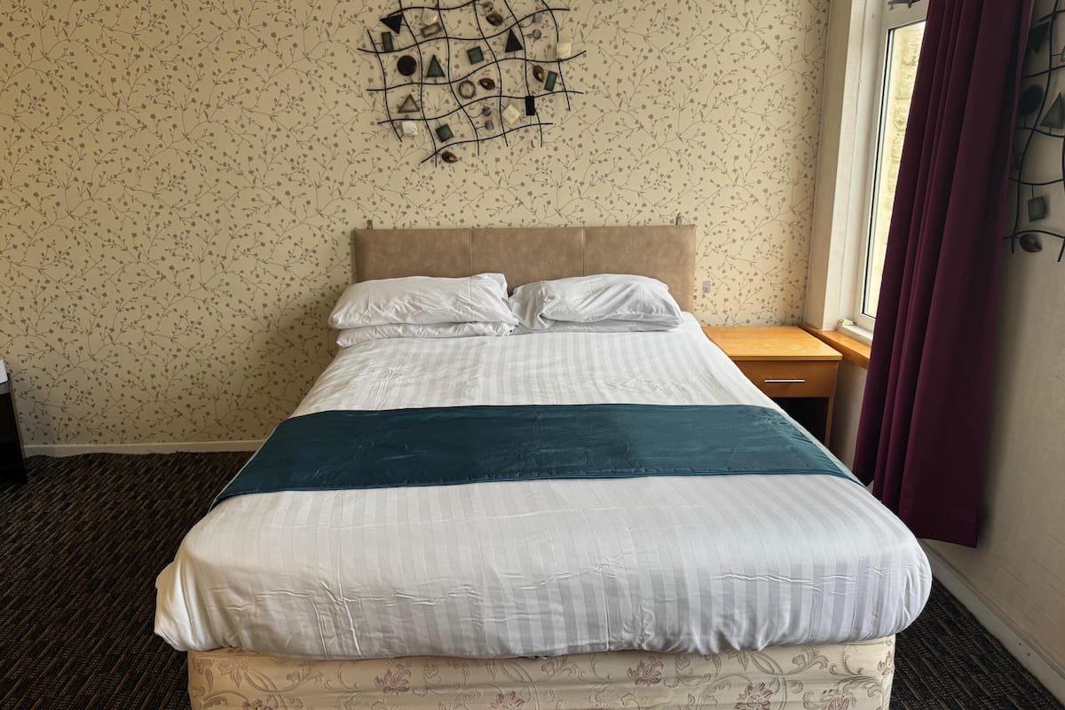 Una cama bien hecha con cabecera, una pieza decorativa en la pared superior y una ventana con cortinas.