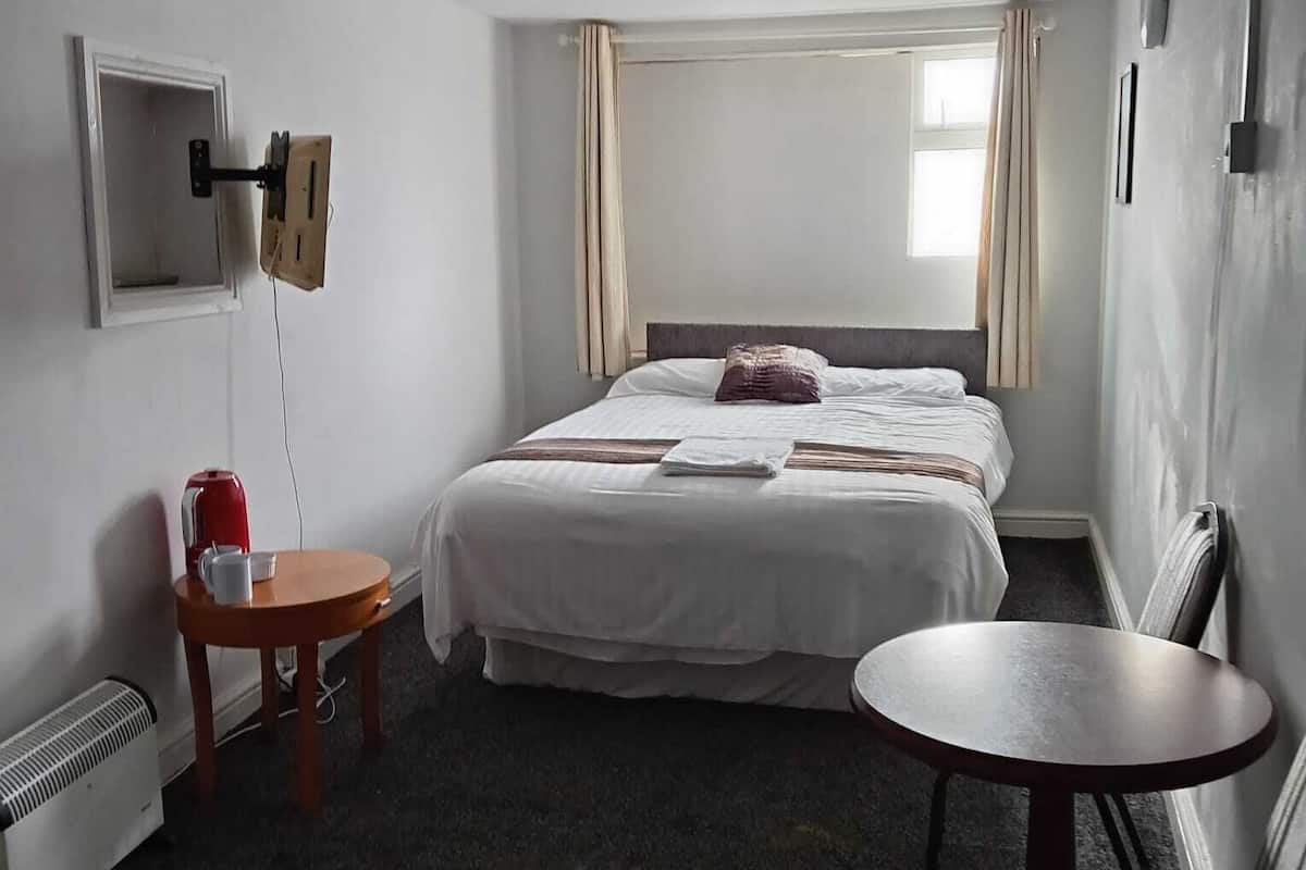 Una habitación de hotel con cama, una mesita, una silla, un calentador y una ventana con cortinas.