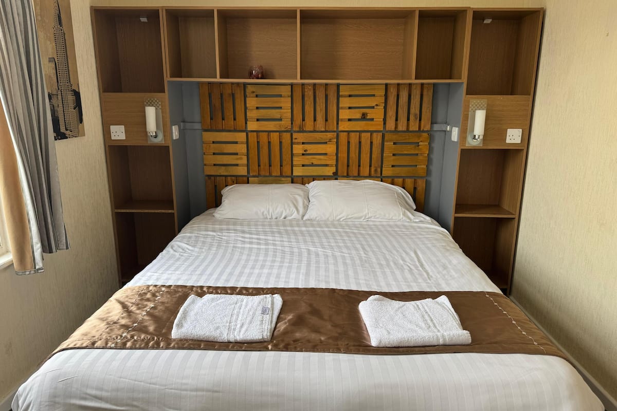 Una habitación de hotel con cabecera de madera, cama con ropa blanca y manta marrón.