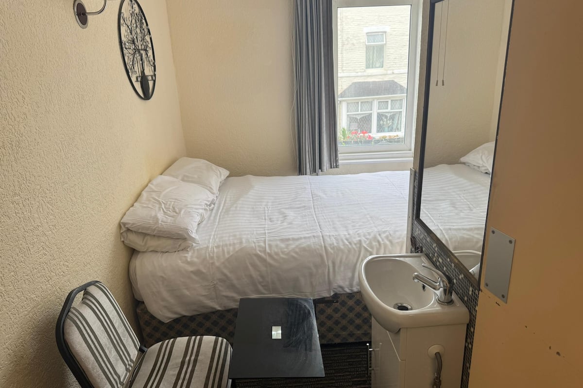 Habitación de hotel con cama, un pequeño escritorio, una silla, un lavamanos y ventana con vista a un edificio.