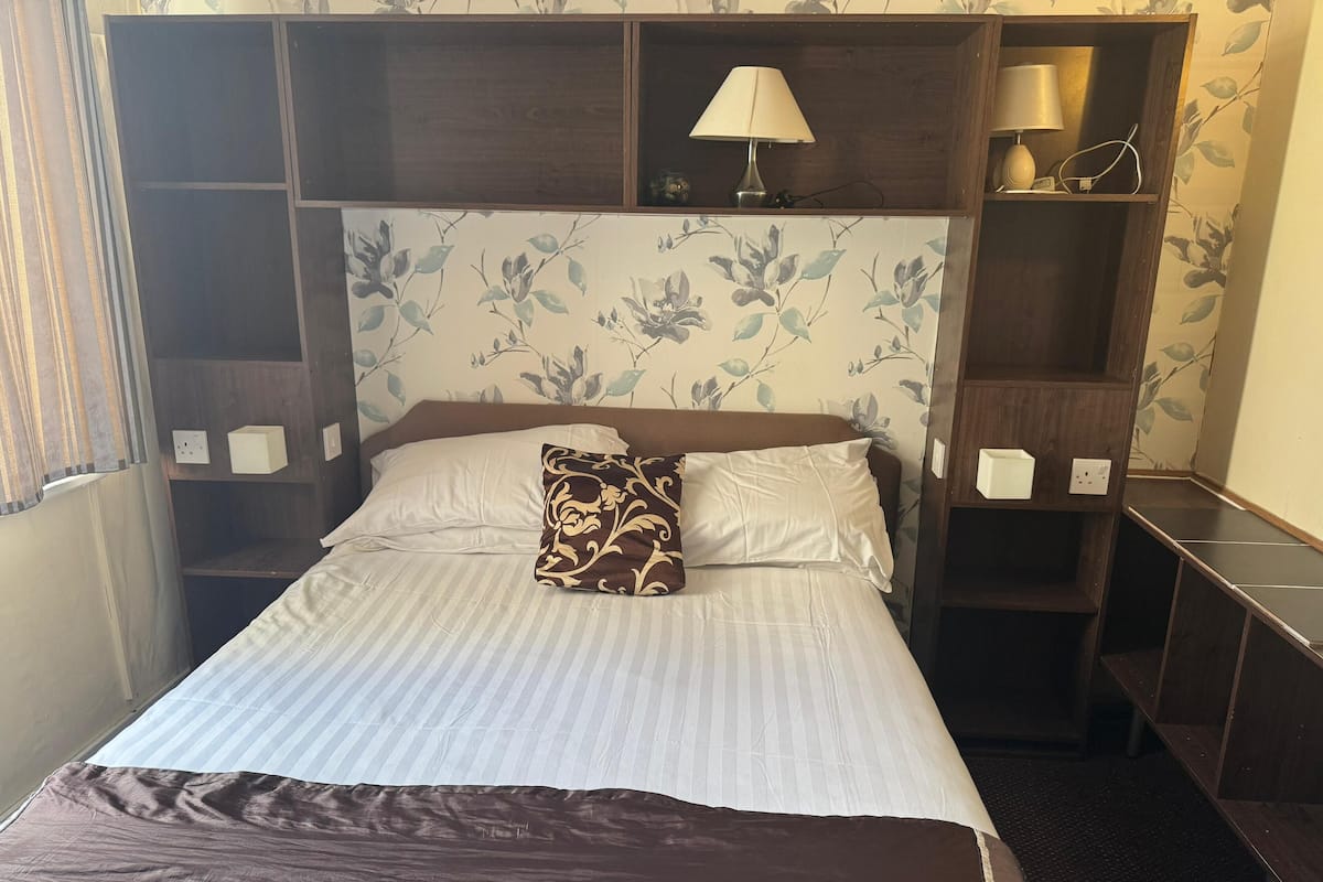 Una cama bien hecha con una colcha a rayas, dos almohadas y un cojín decorativo. Hay una mesita de noche con una lámpara y un enchufe. La habitación tiene un cabecero de madera y un estampado de papel tapiz floral.