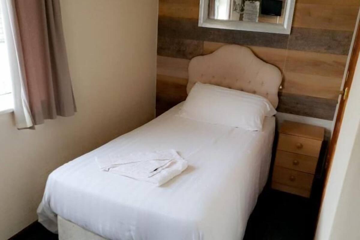 Una cama pequeña y bien hecha en una habitación con cabecera de madera, una ventana con cortinas y una mesita de noche de madera.