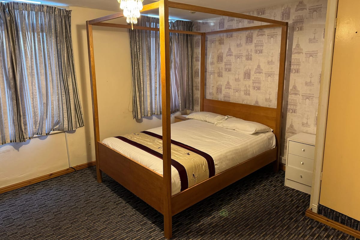 Una cama con dosel de madera y ropa de cama blanca, un candelabro colgante arriba y un papel tapiz con diseños arquitectónicos.