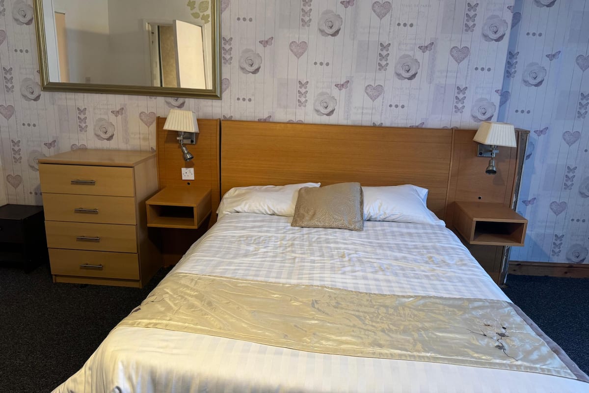 Una habitación de hotel con cama, una mesita de noche de madera con lámpara, un aparador y un espejo en la pared.