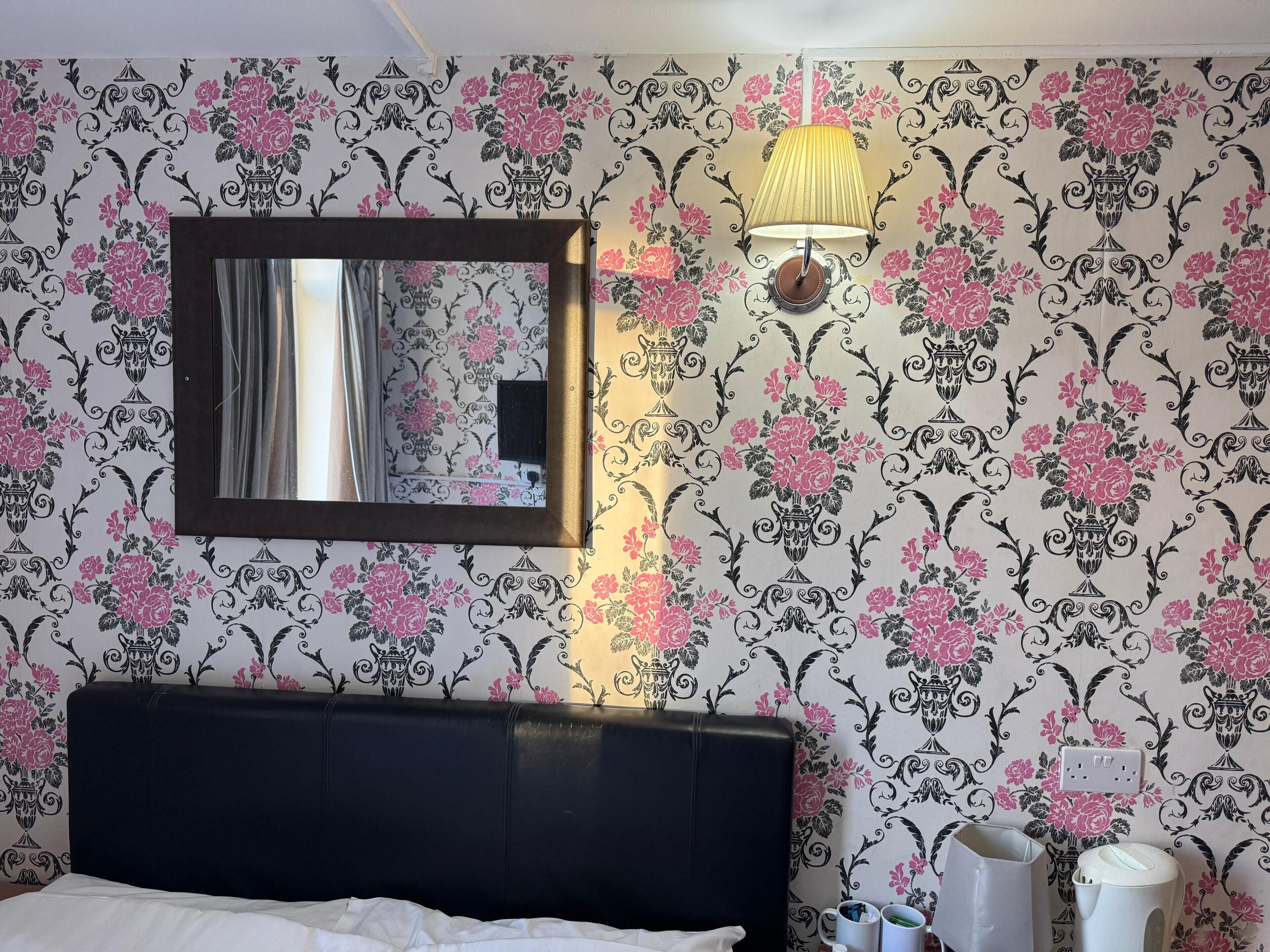 A hotel room with floral wallpaper, a bed with a black headboard, a mirror, a wall-mounted lamp, and a small table with a kettle and cups.