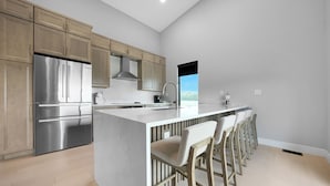 A modern kitchen with stainless steel appliances, a large island with bar stools, and a view of the outdoors. - New Modern Home in the Finger Lakes (Romulus)