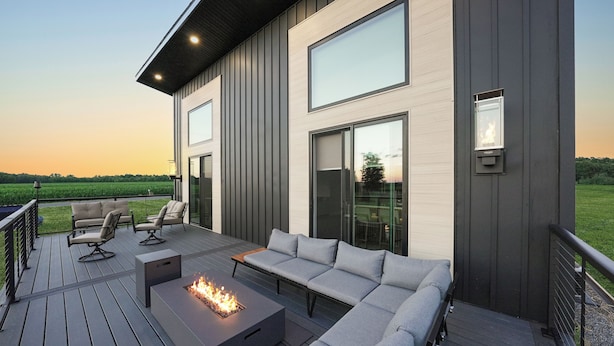 A modern outdoor deck with a fire pit, seating, and large windows overlooking a field. - New Modern Home in the Finger Lakes (Romulus)