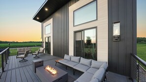 Une terrasse extérieure moderne, dotée d’un foyer, de sièges et de grandes fenêtres donnant sur un champ.