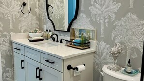 A bathroom with a white vanity, a mirror with a black frame, a toilet, and a blue rug. - 3-bedroom condo in charming Melbourne Beach with AC (Melbourne Beach)