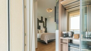 Une chambre d’hôtel moderne avec un plancher en bois, un lit recouvert d’une courtepointe beige, une petite kitchenette équipée d’une machine à café, de verres à vin et d’un panier.