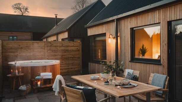 Eine Holzterrasse mit Whirlpool, Esstisch und Stühlen sowie ein großes Fenster mit Blick auf den Garten.