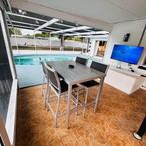 A covered outdoor dining area with a pool, a TV, and a white cabinet.
