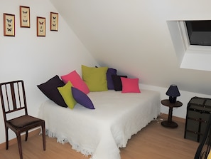 A bedroom with a bed, colorful pillows, a wooden chair, a small table with a lamp, and framed butterfly pictures on the wall.